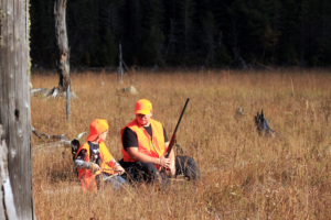 Father and son dressed in orange vest and hats sit on a log during their family hunting trip.