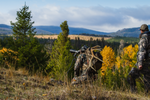 Men dressed in camo with backpacks on a guided hunt on a ridge overlooking pine trees and mountains in the background.